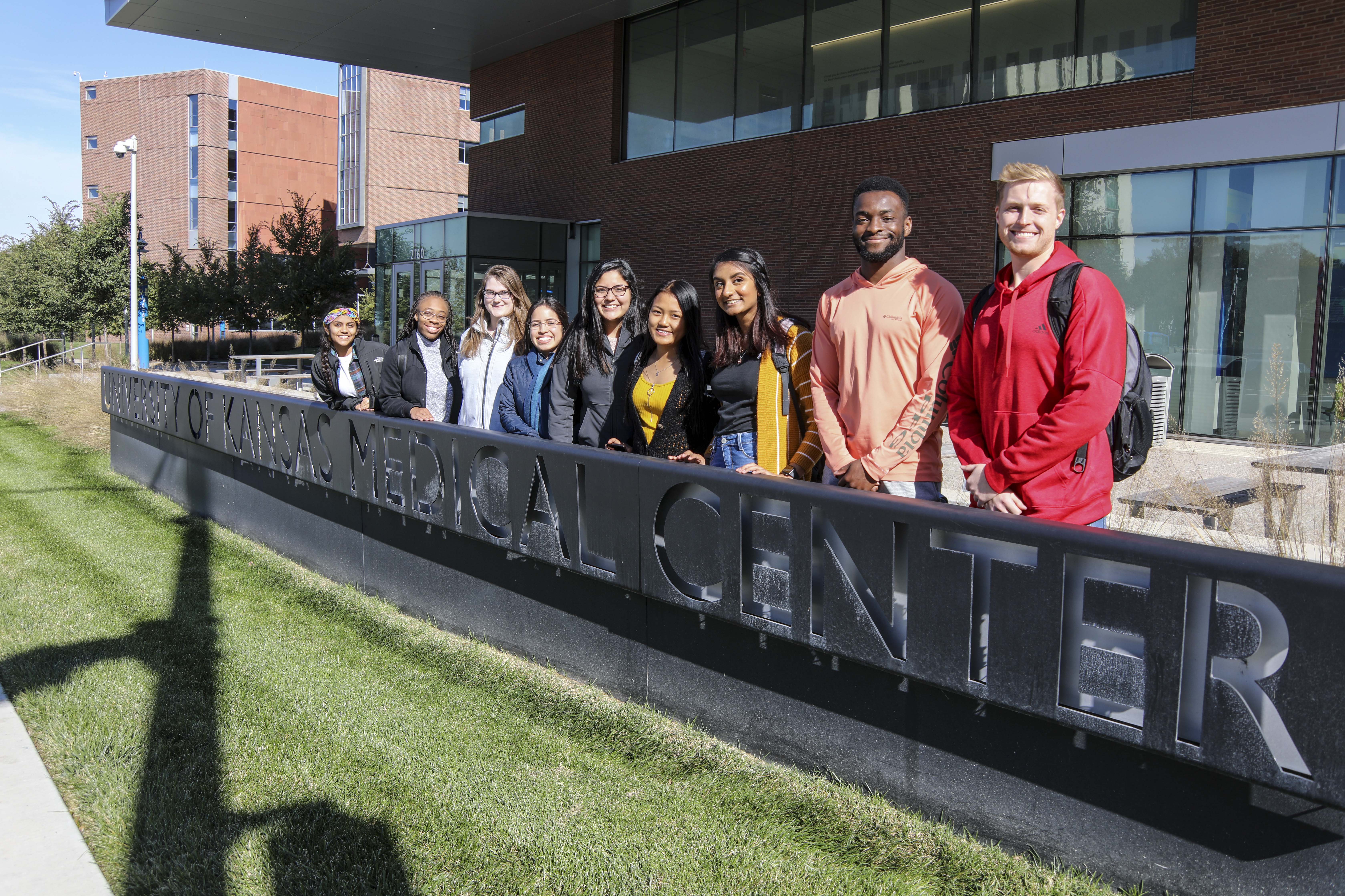 A diverse group of 9 students stand in front of the KU Med Center Sign
